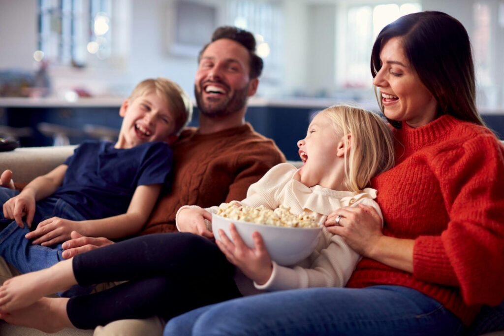 Family Sitting On Sofa With Popcorn Laughing Watching Comedy On TV Together